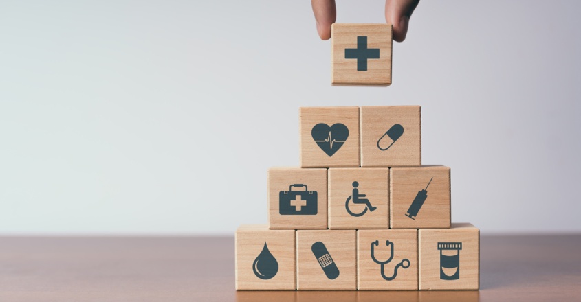 5 druhů zákonného pojištění: Ruka skládající pyramidu z dřevěných kostek, na kterých jsou symboly ze zdravotnictví.
