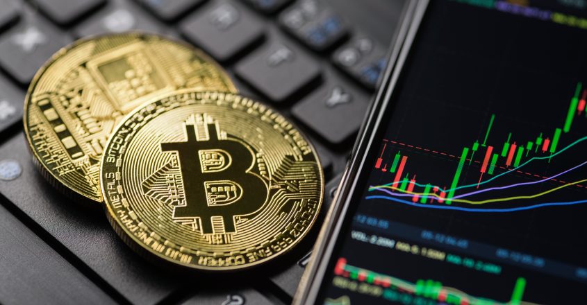 Bitcoiny ležící na klávesnici vedle telefonu s grafem vývoje ceny kryptoměn.