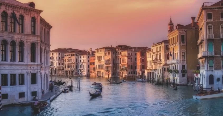Italské Benátky při západu slunce, který ozařuje domy. Na řece plují gondoly.