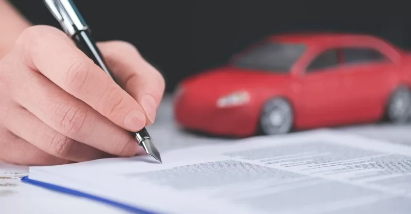 Detail ruky, která perem podepisuje výpověď pojištění auta. V pozadí je červený model vozu.