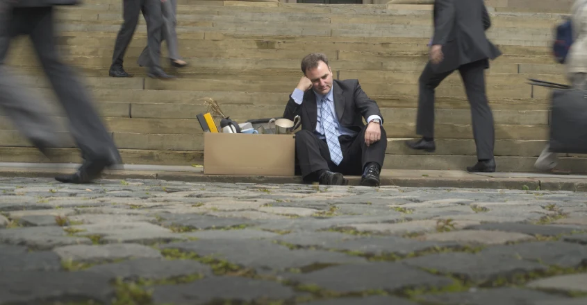 Muž v obleku sedí sklesle na dlážděném povrchu vedle lepenkové krabice s osobními předměty.