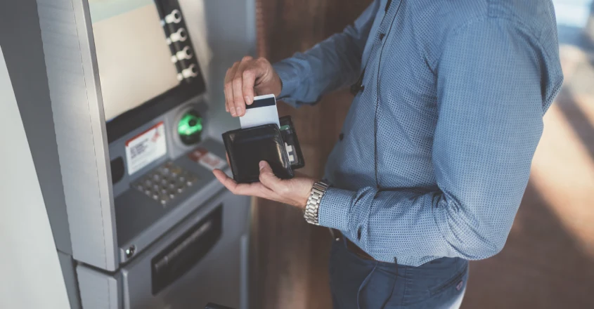 Muž, který stojí u bankomatu a vybírá platební kartu z peněženky.